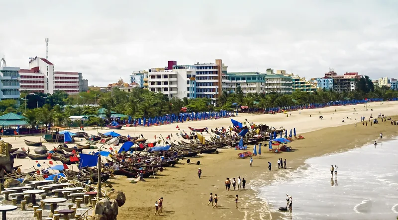 Thuyền đánh cá và những chiếc dù xanh xếp dọc bãi biển Sầm Sơn, phía sau là khách sạn và hàng cây dừa.